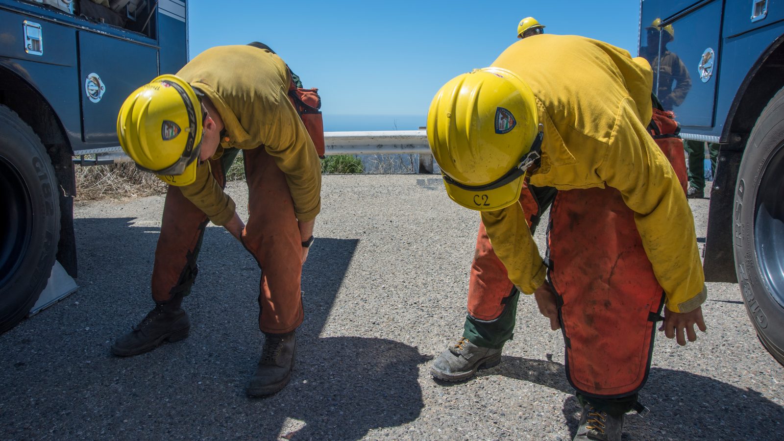 A wildland firefighter tightens the buckle straps of cut-resistant chainsaw chaps over his legs before starting saw work, with crew members doing the same behind him